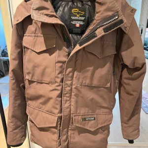 Brun vinterjacka från Canada Goose - Säljer en brun vinterjacka från Canada Goose i mycket bra skick. Jackan har en varm dunfyllning och flera praktiska fickor. Den är långärmad med dragkedja och kardborreknäppning för extra skydd mot kyla. Perfekt för kalla vinterdagar! Små tecken på användning dock ej synligt när jackan är på