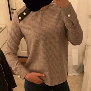 Rutig långärmad topp med knappar - Säljer en stilren rutig topp med långa ärmar och snygga knappdetaljer på axeln och ärmsluten. Perfekt för både vardag och festliga tillfällen. Passar bra till jeans eller kjol för en chic look.
