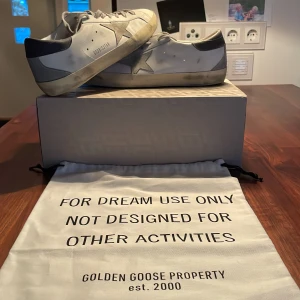 Golden Goose Super-Star sneakers - Säljer ett par Golden Goose Super-Star sneakers i vitt med grå detaljer. De har en cool, sliten look som är typisk för märket. Skorna är i skinn och har snörning. Perfekta för en avslappnad men stilren look. Kommer med originalpåse och låda. Golden goose Passport kommer med samt dust bag. Använde dom ungefär 5 gånger. Nya kostar dom 5500kr. 