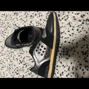 Valentino Garavani sneakers i svart och silver - Snygga Valentino Garavani sneakers i svart och silver med coola nitar på sidorna. Skorna har en stilren design med snörning och en bekväm sula. Perfekta för att ge din outfit en lyxig touch. Använd dem till både vardags och fest!, det är i storlek 43 och är lite använda ingen dust bag eller kartong, fast pris prutat och klart köparen står för frakten.