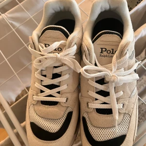 Vita sneakers från Polo Ralph Lauren - Säljer ett par snygga vita sneakers från Polo Ralph Lauren i storlek 45. De har en stilren design med svarta detaljer och är tillverkade i en mix av läder och textil. Perfekta för både vardag och fest! Skorna är i mycket bra skick och har klassisk snörning.