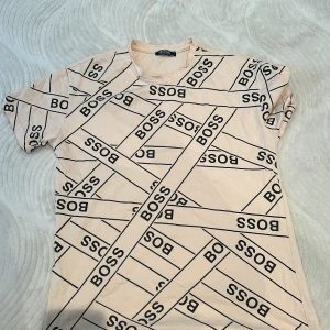 Hugo Boss  - Säljer en beige t-shirt från Boss med ett coolt mönster av loggan över hela framsidan. Den är kortärmad och perfekt för en avslappnad stil. Passar bra till både jeans och shorts. Perfekt för sommardagar eller en chill festkväll.Beige t-shirt från Boss. Det är storlek M men passar även S