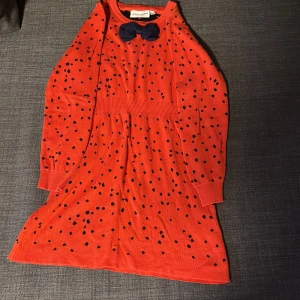 Orange prickig klänning från Mini Rodini - Superfin orange klänning från Mini Rodini med svarta prickar och en söt mörkblå rosett vid halsen. Klänningen är långärmad och har en bekväm passform, perfekt för både lek och fest. Tillverkad i ekologiskt material för extra komfort.