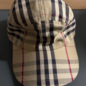 Äkta Burberry keps (vintage) - Snygg rutig keps från Burberry i beige, svart och röd. Perfekt för att ge din outfit en klassisk touch. Tillverkad i ett slitstarkt material och har en justerbar passform. Passar både till vardags och mer uppklädda tillfällen.