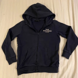 Mörkblå hoodie från Peak Performance - Säljer en snygg mörkblå hoodie från Peak Performance i bra skick. Den har en dragkedja framtill och en broderad logga på bröstet. Perfekt för höst och vår, med långa ärmar och en mysig huva. Passar både till vardags och träning!