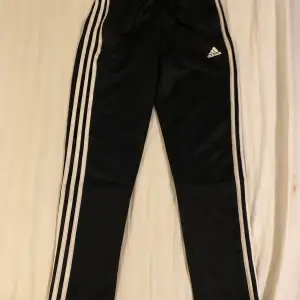 Säljer ett par svarta träningsbyxor från Adidas i storlek 150 . Byxorna har de klassiska tre vita ränderna längs sidorna och Adidas-loggan på framsidan. Perfekta för träning eller en avslappnad stil. Materialet är mjukt och bekvämt, idealiskt för både inomhus och utomhusaktiviteter.