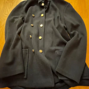 Svart kappa från Petri - Säljer en elegant svart kappa från Petri i storlek 40. Den har en klassisk design med dubbla knapprader och långa ärmar. Perfekt för höst och vinter, och ger en stilren look. Kappan är i bra skick och redo för nya äventyr!