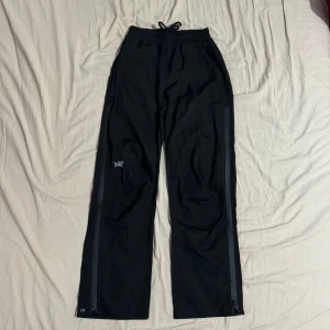 Svarta byxor från Arc’teryx - Säljer ett par svarta byxor i mycket bra skick. De har en broderad detalj på sidan och är högmidjade med en loose passform. Perfekta för höst och vår, och de har praktiska dragkedjor längs benen. Materialet känns slitstarkt och bekvämt.