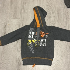 Grå hoodie med motorcykeltryck - Säljer en cool grå hoodie med motorcykeltryck i gult och orange. Perfekt för små äventyrare! Tröjan har en dragkedja framtill och en mysig huva med orange foder och är i bra skick 