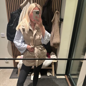 Beige dunväst med knyt - Säljer en snygg beige dunväst med knyt i midjan. Perfekt för höst och vår, den har en normal passform och är superbekväm. Västen har en dragkedja framtill och är i mycket bra skick. Passar perfekt över en tröja eller skjorta för en stilren look.