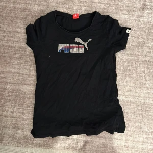 Svart t-shirt från Puma - Säljer en svart t-shirt från Puma i bra skick. Den har ett coolt Puma-tryck i flera färger på bröstet. Perfekt för vardagsbruk eller träning. T-shirten är kortärmad och har en normal passform. Skön och stilren design! Säljer för att jag inte andvänder den