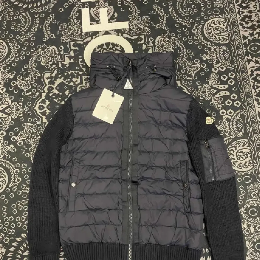 Säljer en snygg svart jacka från Moncler. Den har en quiltad design med stickade ärmar och en praktisk huva. Perfekt för höst och vinter! Jackan har dragkedja och en liten logga på ärmen. Passar både till vardags och lite finare tillfällen.. Jackor.