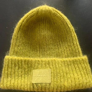 Limegrön stickad mössa från Carin Wester ull/ mohair  - Säljer en superfin gul stickad mössa från Carin Wester. Den är ribbad och har en snygg logga framtill. Perfekt för att hålla sig varm och stilren under höst och vinter. Passar till alla outfits och ger en färgklick i vardagen! Ull/ mohair.