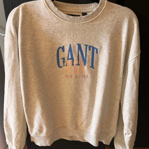 Grå sweatshirt från GANT - Säljer en snygg grå sweatshirt från GANT med tryck i blått och rött. Tröjan har en klassisk rund hals och långa ärmar, perfekt för en avslappnad stil. Den är i bra skick och passar perfekt för både höst och vår. Perfekt för dig som vill ha en stilren och bekväm look!