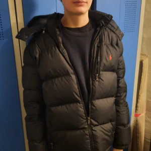 Svart dunjacka från Polo Ralph Lauren - Säljer en svart dunjacka från Polo Ralph Lauren i mycket bra skick. Jackan har en dragkedja och är perfekt för vintern. Den är långärmad och har en klassisk pufferstil som håller dig varm och snygg. Perfekt för kalla dagar! 