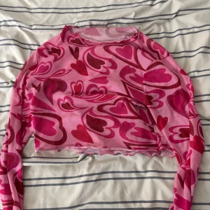 Rosa långärmad topp med hjärtmönster - Säljer en superfin rosa långärmad topp med ett coolt hjärtmönster i olika nyanser av rosa och rött. Toppen är i ett stretchigt material och har en croppad passform. Kan passar som storlek M också. 
