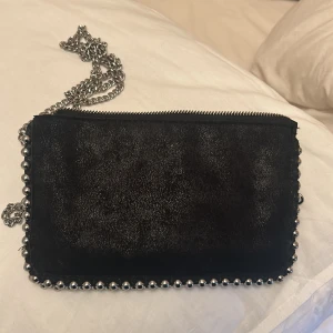 Svart axelväska med kedja - Snygg svart axelväska i mocka med coola silverdetaljer. Väskan har en kedja som axelrem och en dragkedja upptill. Perfekt för en utekväll eller fest! Den är i mycket bra skick och har en stilren design med små silverkulor längs kanterna.