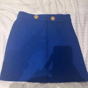 Blå kjol med guldknappar - Säljer en superfin blå kjol med två dekorativa guldknappar i midjan. Kjolen har en klassisk och stilren design som passar perfekt för både vardag och fest. Den är i mycket bra skick och har en normal passform. Perfekt för höst och vår!