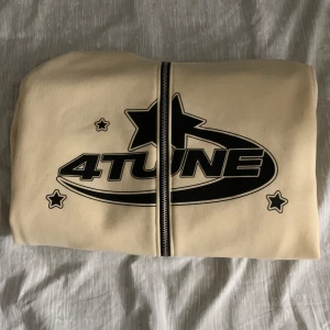 Beige hoodie med tryck från 4Tune - Säljer en snygg beige hoodie från 4Tune med coolt svart tryck fram och bak. Den har en dragkedja och en mysig huva, perfekt för kyliga dagar. Fickor framtill för extra stil och funktionalitet. Passar perfekt för en avslappnad look!