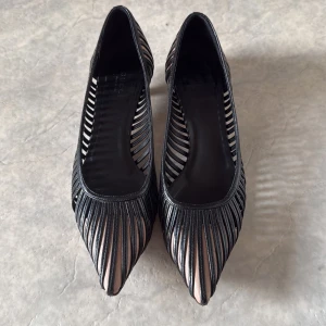 Svarta klackskor från Gucci - Säljer ett par eleganta svarta klackskor från Gucci i bra skick. Skorna har en unik design med remmar och spetsig tå, vilket ger dem en stilren look. Perfekta för festliga tillfällen eller en utekväll. Lite slitage på sulorna, men annars i fint skick.