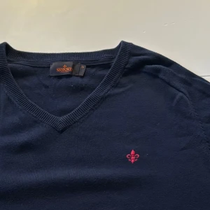 Mörkblå tröja från Morris - Säljer en snygg mörkblå tröja från Morris i storlek M. Den har en v-ringad design och ett litet rött broderat emblem på bröstet. Perfekt för både vardag och festliga tillfällen. Tröjan är i mycket bra skick och passar perfekt till höst och vinter. Hör av dig vid intresse!