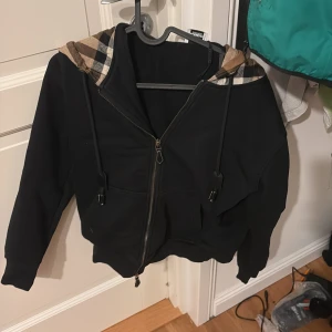 Svart hoodie från Burberry - Säljer en snygg svart hoodie från Burberry med klassiskt rutigt mönster i huvan. Den har dragkedja framtill och långa ärmar. Perfekt för höst och vår när du vill vara både bekväm och stilren. Passar till både vardag och fest. Ny pris :7000 