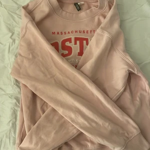 Rosa sweatshirt från H&M Divided - Säljer en superfin rosa sweatshirt från H&M Divided med tryck av 'Boston Massachusetts' i rött och vitt. Tröjan har långa ärmar och ribbade muddar vid ärmslut och nederkant. Perfekt för en avslappnad stil eller en mysig dag hemma. 🩷