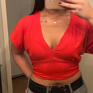 Röd kortärmad topp med knytning i ryggen - Säljer en snygg röd topp med korta ärmar och v-ringning. Toppen är croppad och har en öppen rygg med knytning, vilket ger en cool och avslappnad stil. Perfekt för sommardagar eller en utekväll. 