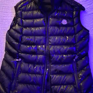 Säljer en snygg svart dunväst från Moncler i mycket bra skick. Västen har en glansig finish och är perfekt för höst och vinter.  två praktiska fickor med dragkedjor. Moncler-loggan sitter på bröstet. Perfekt för att hålla sig varm och stilren!                                                                        Enda nackdelen är att dragkedjan framtill har gått av och att cartoonen har tappat färg 