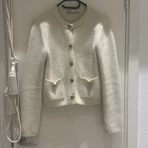Vit kavaj från Zara - Säljer en elegant vit kavaj från Zara i mycket bra skick. Den har en fin struktur och är dekorerad med guldknappar framtill. Kavajen har långa ärmar och två fickor med dekorativa detaljer. Perfekt för höst och vår!