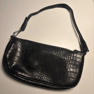 Svart axelväska - Svart axelväska  i läderimitation med krokodilmönster. Väskan har en dragkedja upptill och har två små små fack på insidan. Köpt second hand och tyvärr ingen lapp så vet ej vart den är ifrån. Använd ett fåtal gånger☺️🫶