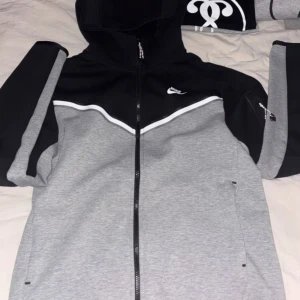 Grå/svart  huvtröja från Nike - Säljer en snygg grå och svart huvtröja från Nike med dragkedja framtill. Tröjan har en cool design med vita detaljer och en praktisk huva. Perfekt för en sportig look!