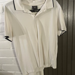 Vit pikétröja från H&M - Säljer en stilren vit pikétröja från H&M i slim fit. Tröjan har korta ärmar och svarta detaljer på kragen och ärmsluten. Perfekt för en avslappnad men snygg look.