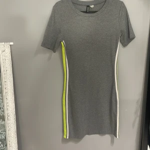 Grå klänning med neongula detaljer - Trendig grå klänning med neongula och vita ränder längs sidorna. Den har en kort ärm och en kroppsnära passform som ger en sportig look. Perfekt för en avslappnad stil.