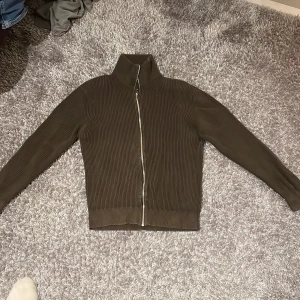 Brun ribbad stickad tröja  från East West - Säljer en brun stickad tröja från East West i storlek M. Jackan har en dragkedja framtill och en hög krage. Perfekt för en stilren look.