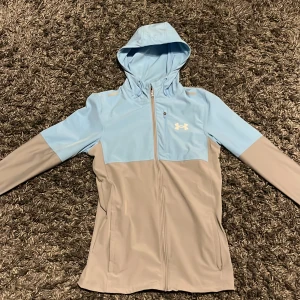 Blå och grå Windbreaker från Under Armour - Snygg blå och grå jacka från Under Armour med huva och dragkedja. Perfekt för träning eller en aktiv dag ute. Jackan har en sportig look med långärmade ärmar och en bekväm passform.