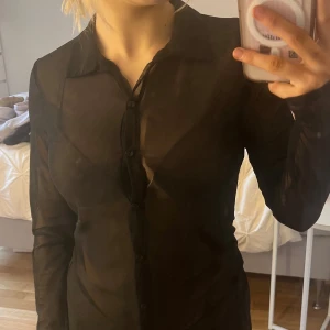 Svart transparent blus - Snygg svart transparent blus med knappar framtill. Perfekt för en stilren look. Blusen har långa ärmar och en klassisk krage. Genomskinlig. 