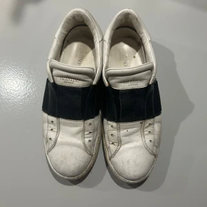 Valentino Garavani sneakers i vitt och svart - Snygga Valentino Garavani sneakers i vitt skinn med en bred svart rem över foten. Skorna har en klassisk design med rund tå och perforerade detaljer. Perfekta för en stilren look. Skick 7/10 pris är diskuterbart. Storlek 42 passar 43.