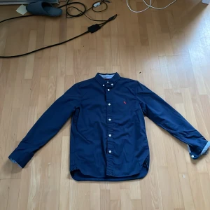 Mörkblå skjorta från L.O.G.G. - Snygg mörkblå skjorta från L.O.G.G. med klassisk krage och knappar framtill. Perfekt för en stilren look. Skjortan har långa ärmar och en liten broderad detalj på bröstet.