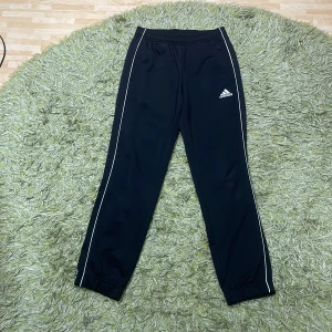 Svarta träningsbyxor från Adidas - Snygga svarta träningsbyxor från Adidas med vita detaljer längs sidorna. Byxorna har en elastisk midja och är perfekta för sport och träning. Adidas-loggan är tryckt på ena benet.