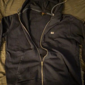 Mörkblå hoodie från Lee - Säljer en mörkblå hoodie från Lee med dragkedja och justerbar huva. Tröjan har en broderad logotyp på bröstet och är perfekt för en avslappnad stil. Den har långa ärmar och en klassisk passform. Perfekt för kyliga dagar! Mycket bra skick storlek L