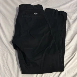 Svarta byxor från Dickies - Säljer ett par klassiska svarta byxor från Dickies. De har en rak passform och är tillverkade i en blandning av polyester och bomull. Perfekta för en stilren look. Byxorna har en diskret logga på bakfickan och stängs med dragkedja och knapp.