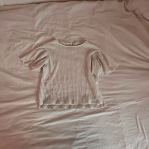 Vit topp från Zara - Säljer en söt vit topp från Zara i ett krinklat material. Toppen har korta ärmar med volangdetaljer och en liten knapp i nacken. Perfekt för en avslappnad stil.