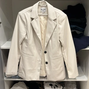 Vit kavaj från NA-KD - Snygg vit kavaj från NA-KD med klassisk design och två knappar framtill. Perfekt för en stilren look. Tillverkad i ett syntetmaterial som ger en elegant finish. Passar bra till både jeans och kjol.