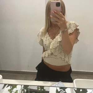 Beige spetsblus med volanger - Charmig beige blus i spets med volanger och v-ringning. Perfekt för en somrig look med sin luftiga design och detaljerade spetsmönster. Blusen är croppad och har en romantisk stil.