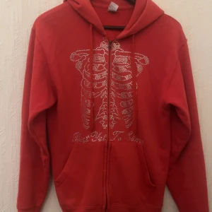 Röd hoodie - Cool röd hoodie med dragkedja och ett glittrigt skelettmönster på framsidan. Perfekt för att sticka ut med en unik stil. Den har en klassisk passform med långa ärmar och en bekväm huva.