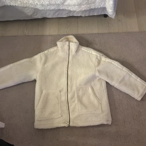 Vit teddyjacka med dragkedja - Mysig vit teddyjacka med dragkedja och två stora fickor framtill. Perfekt för kyliga dagar när du vill hålla dig varm och bekväm. Jackan har en hög krage och långa ärmar.