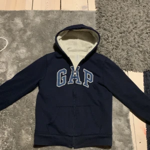 Mörkblå hoodie från GAP - Säljer en mörkblå hoodie från GAP med dragkedja och vit fleecefoder i luvan. Perfekt för kyliga dagar med sin mysiga insida. GAP-loggan är tryckt på framsidan blåt och vit 