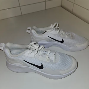 Vita sneakers från Nike - Säljer ett par vita Nike sneakers med svart swoosh. Skorna har snörning och är i syntetmaterial. Perfekta för en sportig look.
