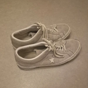Grå sneakers från Converse - Säljer ett par stilrena grå sneakers från Converse med klassisk stjärndetalj på sidan. Skorna har snörning och är tillverkade i mocka för en trendig look. Perfekta för en avslappnad stil.
